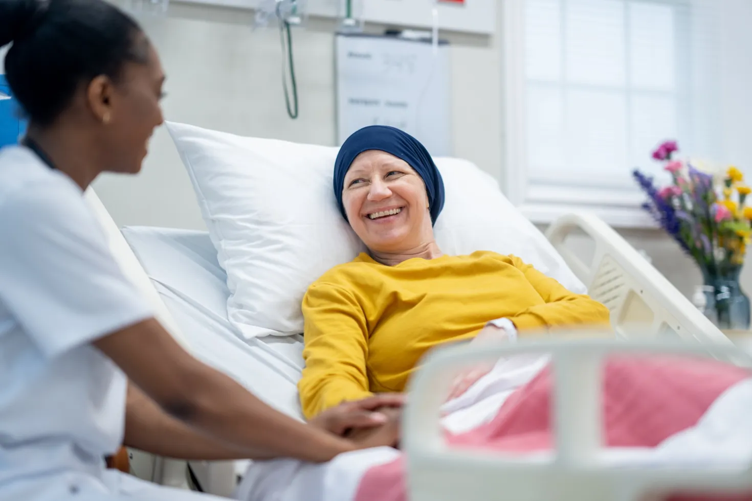 A female nurse of African decent sits at the bedside of a mature female Oncology patient as she checks in on her during rounds. The patient is dressed comfortably and has a head scarf on to keep warm.