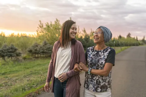 A senior woman with cancer walks with her adult daughter at sunset down a rural road. The affectionate pair are talking and walking with arms linked.